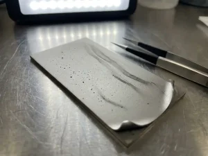 Präzisionsbeschichtete Metalloberfläche mit Dünnschichtdefekten, Nadelstichporen und Kantenablösung unter Laborlicht, Reinraumpinzette daneben.