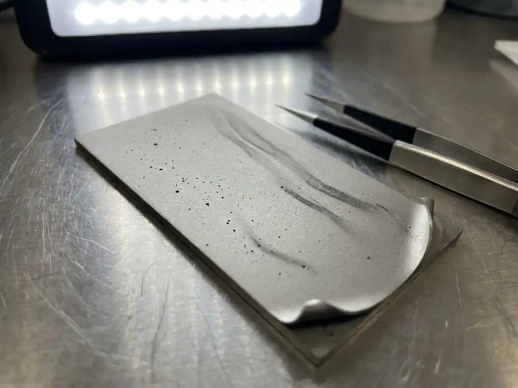 Präzisionsbeschichtete Metalloberfläche mit Dünnschichtdefekten, Nadelstichporen und Kantenablösung unter Laborlicht, Reinraumpinzette daneben.