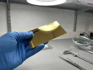 Behandschuhte Hand hält dünnes Edelmetallsubstrat ins Licht, spiegelglatte Beschichtung sichtbar, Präzisionsprüfstand im Hintergrund.