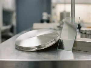 Poliertes Palladium-Sputtertarget auf Stahloberfläche neben irisierendem dünnschichtbeschichtetem Glassubstrat im Labor.