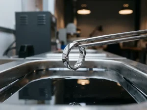 Silberner Ring wird mit Metallzange aus einem Rhodium-Galvanikbad gehoben, spiegelnde Oberfläche im professionellen Schmuckworkshop.