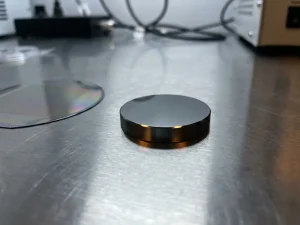 Ruthenium-Sputtertarget-Scheibe neben einem siliziumwafer mit irisierendem Dünnfilm auf polierter Stahloberfläche.